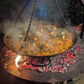 Stoom stijgt op van een verse Paella Valenciana in een grote authentieke pan, sfeervol bereid op een feloranje gloeiend houtvuur in de donkere avondlucht.